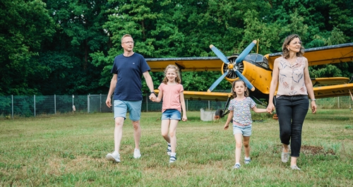 Mutter, Vater, zwei Töchter laufen Hand in Hand über eine grüne Wiese vor einem historischen Doppeldecker Flugzeug